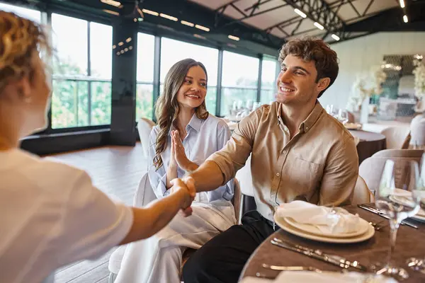 Sonriente hombre estrechando la mano con gerente de eventos cerca de novia muy alegre en el lugar moderno de la boda - foto de stock