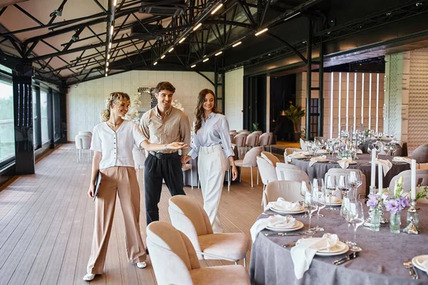 Event manager showing festive tables to happy couple in love in event hall with wedding decor — Stock Photo