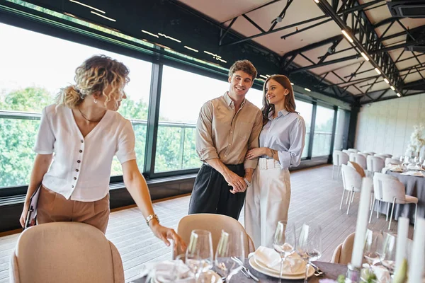 Alegre pareja enamorada cerca de gestor de eventos y mesas con ambiente festivo en la moderna sala de banquetes - foto de stock