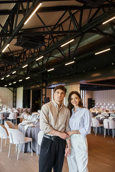 Casal alegre no amor olhando para a câmera perto da mesa festiva no salão de eventos, preparação do casamento — Fotografia de Stock