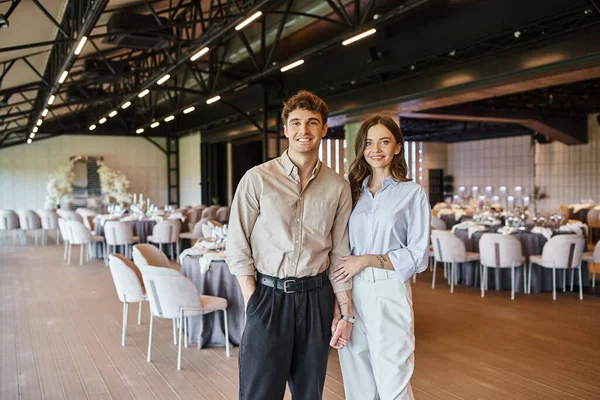 Joyeux couple amoureux souriant à la caméra près de la table de fête dans le lieu de mariage, cadre romantique — Photo de stock