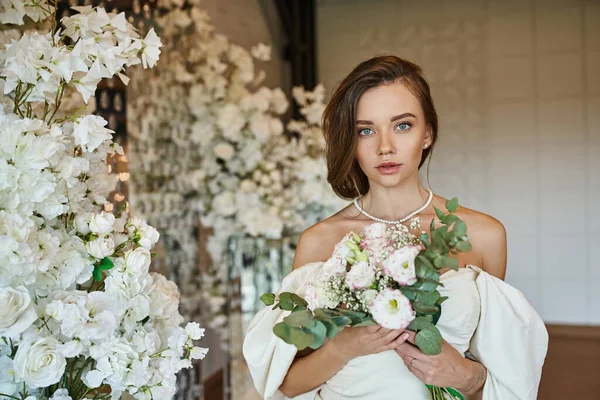 Mulher elegante em vestido de noiva branco com buquê de noiva olhando para a câmera perto de decoração floral festiva — Fotografia de Stock