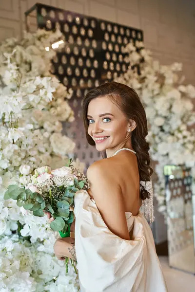 Joyful woman in white wedding dress with bridal bouquet smiling at camera near festive floral decor — Stock Photo