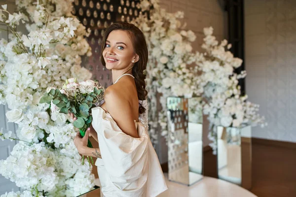 Heureuse jeune femme en robe de mariée posant avec bouquet de mariée près de la décoration florale dans la salle d'événements — Photo de stock