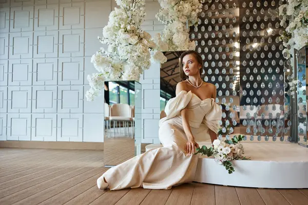 Piena lunghezza della sposa in abito da sposa elegante seduto vicino a decorazioni floreali bianche nella sala banchetti — Foto stock