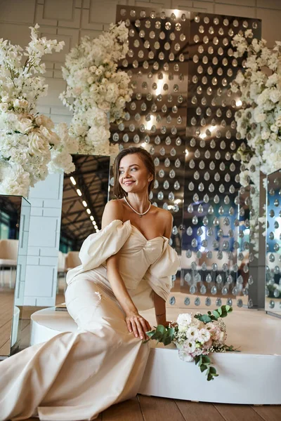 Joyeuse jeune mariée en robe de mariée blanche assise près de décor floral blanc dans le hall de célébration — Photo de stock