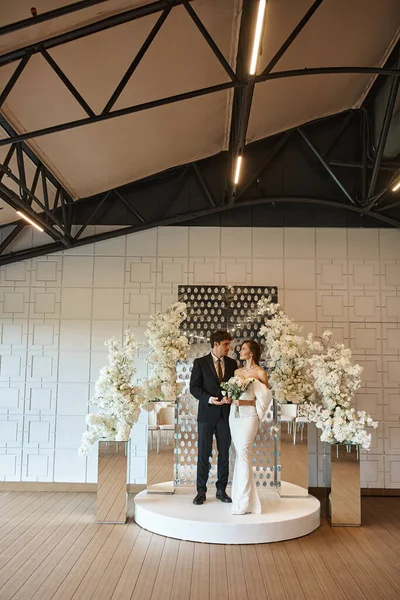 Comprimento total do casal recém-casado elegante no local do casamento decorado com flores brancas florescendo — Fotografia de Stock