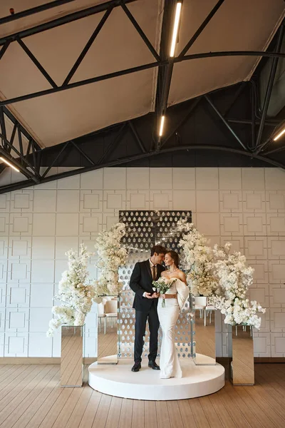 Повна довжина елегантних молодят у весільному залі, прикрашеному білими квітами, особливий день — стокове фото