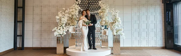 Comprimento total de recém-casados elegantes posando no salão de eventos decorado com flores brancas florescendo, banner — Fotografia de Stock