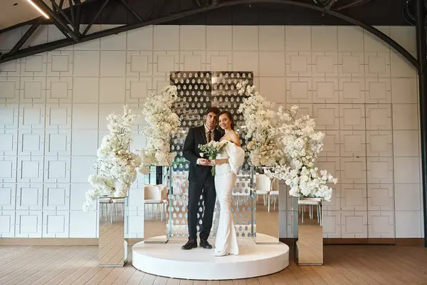 Full length of romantic newlywed couple posing in event hall decorated with white blooming flowers — Stock Photo