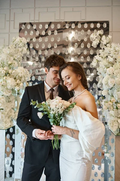 Casal alegre em traje de casamento com buquê de noiva no salão de eventos decorado com flores brancas — Fotografia de Stock