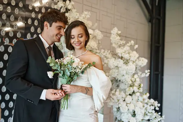 Noivo feliz segurando a mão de noiva encantadora com buquê de noiva perto de decoração floral no salão de eventos — Fotografia de Stock