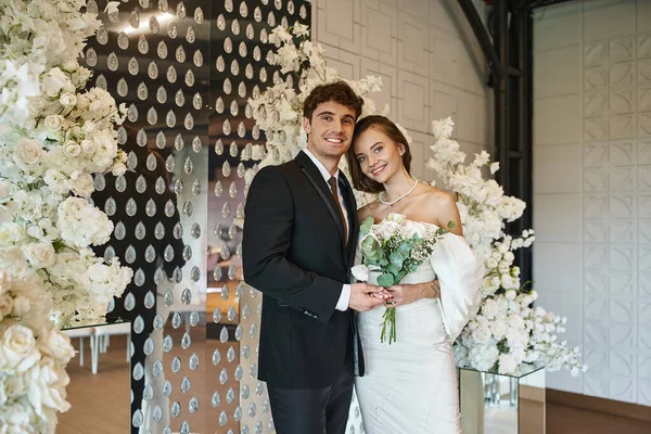 Happy and elegant newlyweds looking at camera in wedding hall decorated with white blooming flowers — Stock Photo