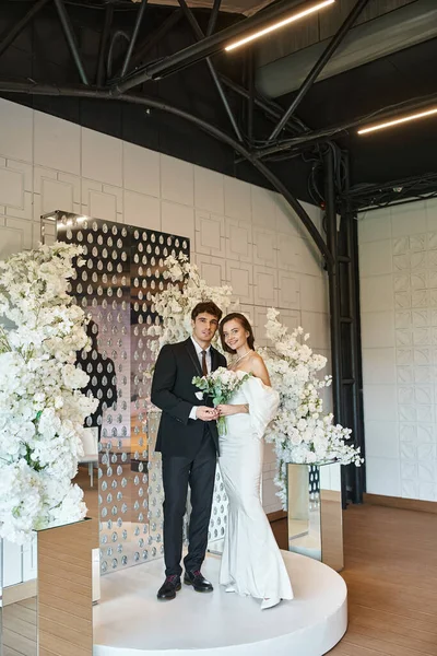 Full length of romantic newlywed couple posing in event hall decorated with white blooming flowers — Stock Photo