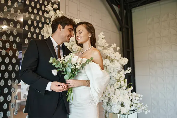 Homem feliz segurando a mão da noiva encantadora com buquê de noiva perto de decoração floral branca no salão de eventos — Fotografia de Stock