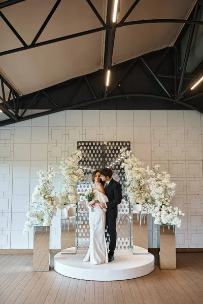 Comprimento total do noivo abraçando mulher encantadora com buquê de noiva perto de decoração floral no salão do banquete — Fotografia de Stock