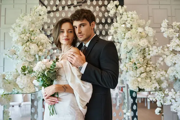 Jovens e elegantes recém-casados olhando para a câmera perto de composição floral branca no local do casamento — Fotografia de Stock