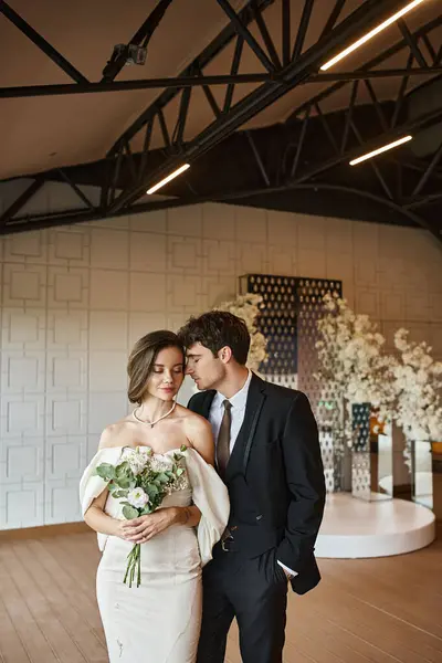 Encantadora novia en vestido de novia blanco cerca del novio en traje negro en salón de eventos con decoración floral - foto de stock