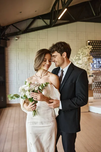 Feliz y encantadora novia con ramo de boda sonriendo cerca del novio elegante en la sala de eventos decorada - foto de stock