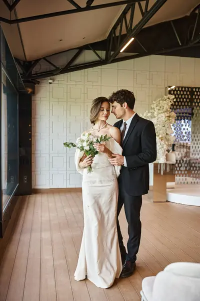 Larga duración de la joven pareja elegante en traje de novia en salón de banquetes moderno con decoración floral - foto de stock