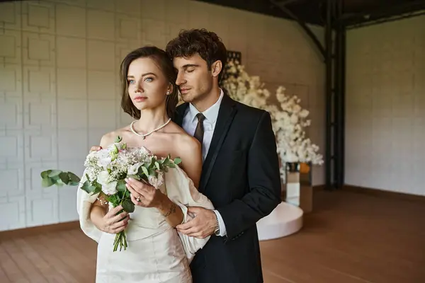 Felice uomo con gli occhi chiusi vicino affascinante sposa in piedi con bouquet da sposa in sala eventi — Foto stock