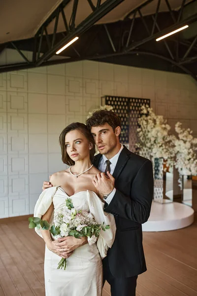 Noivo elegante abraçando noiva encantadora com buquê de casamento e olhando para a câmera no salão de eventos — Fotografia de Stock
