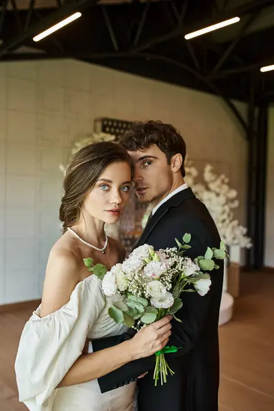 Elegant and romantic newlyweds looking at camera near wedding bouquet in modern banquet hall — Stock Photo