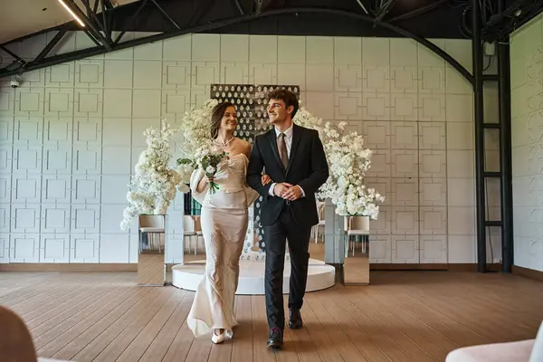 Comprimento total de casal alegre elegante em traje de casamento andando no salão de eventos com decoração floral — Fotografia de Stock