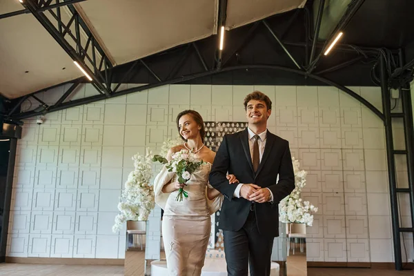Cheerful elegant couple in wedding attire walking in modern event hall with white floral decor — Stock Photo