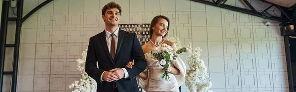 Glückliches Brautpaar in Hochzeitskleidung in moderner Veranstaltungshalle mit weißem Blumendekor, Banner — Stockfoto