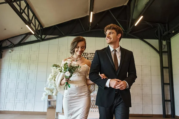 Noiva feliz em vestido branco e noivo em terno preto sorrindo no moderno local de casamento decorado — Fotografia de Stock