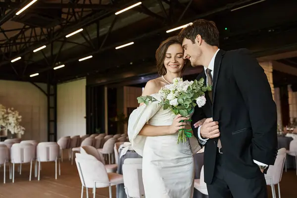 Novio feliz sonriendo con los ojos cerrados cerca de novia encantadora con ramo de boda en la sala de banquetes moderna - foto de stock