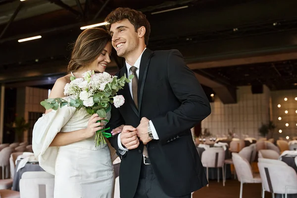 Noivo feliz sorrindo perto de jovem noiva encantadora com buquê de casamento no salão de banquetes moderno — Fotografia de Stock