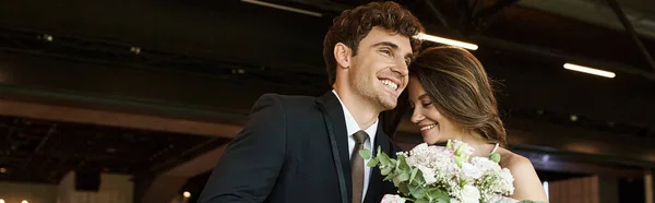 Novio complacido sonriendo cerca de la novia elegante joven con ramo de boda en la sala de banquetes moderna - foto de stock