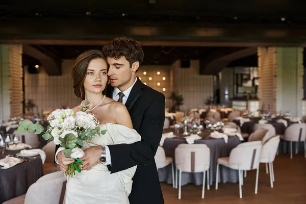 Элегантный мужчина с закрытыми глазами обнимает молодую невесту свадебным букетом в современном зале для мероприятий — стоковое фото