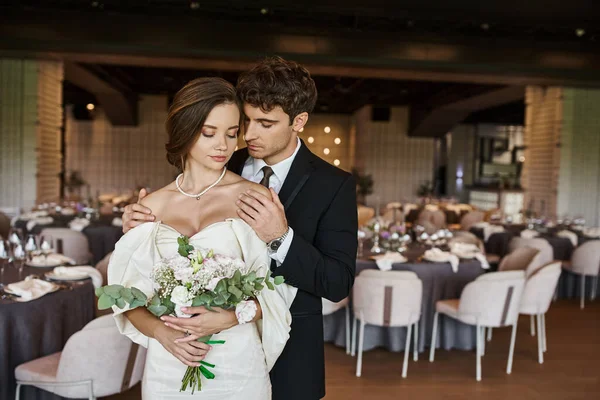 Elegante novio con los ojos cerrados abrazando encantadora novia con ramo de boda en la sala de eventos moderna - foto de stock