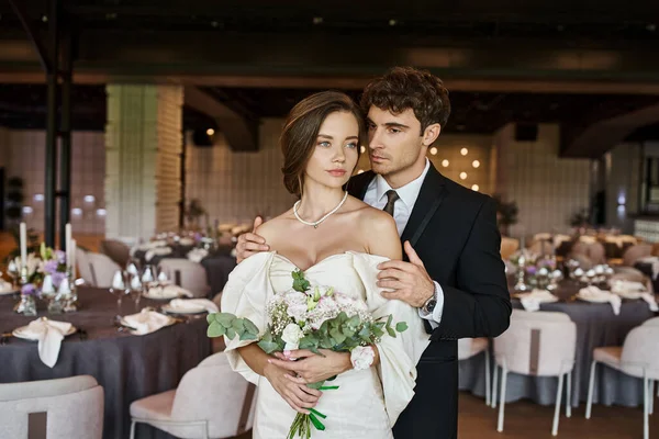 Eleganter Bräutigam umarmt junge und charmante Braut mit Brautstrauß im modernen Bankettsaal — Stockfoto