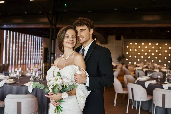 Feliz joven pareja en elegante traje de novia mirando a la cámara en la sala de celebración decorada - foto de stock