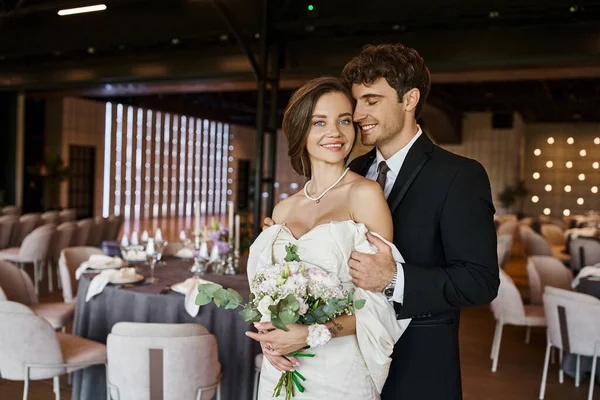 Jovem casal alegre em traje de casamento elegante sorrindo para a câmera no salão de celebração decorado — Fotografia de Stock
