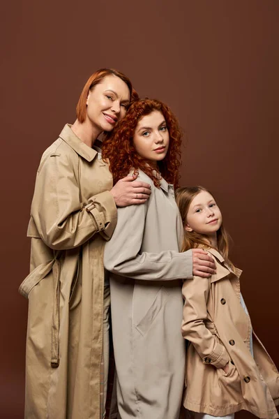 Tres generaciones, mujeres pelirroja feliz y linda chica en abrigos de trinchera posando sobre fondo marrón - foto de stock