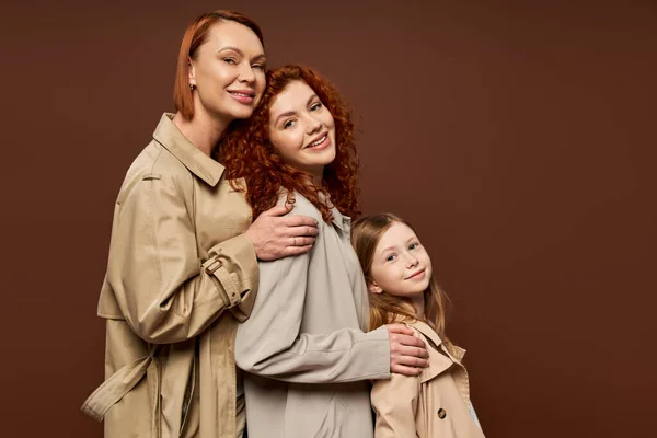 Три покоління, веселі руді жінки і мила дівчина в траншеї пальто позує на коричневому фоні — стокове фото