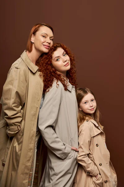 Три поколения, веселые рыжие женщины и девушка в осенних пальто позируя на коричневом фоне — стоковое фото