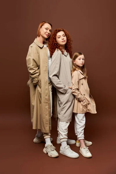 Tres generaciones, mujeres pelirrojas y niñas posando en abrigos sobre fondo marrón, atuendo otoñal - foto de stock
