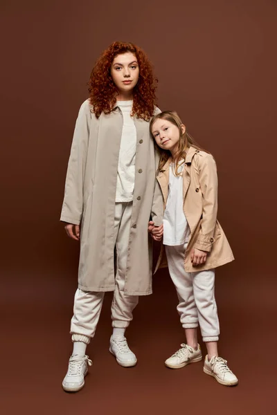 Dos generaciones, madre rizada cogida de la mano con su hija y de pie en abrigos sobre fondo marrón - foto de stock