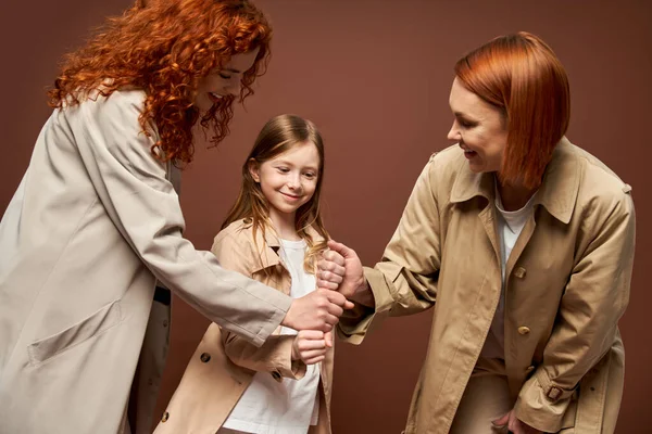 Generations, happy redhead family in coats stacking hands together on brown backdrop, women and girl — Stock Photo