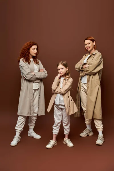Three generations, women in coats posing and looking at girl on brown backdrop, redhead family — Stock Photo