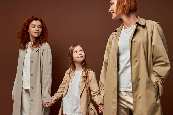 Three generations, women in coats holding hands with happy girl on brown backdrop, redhead family — Stock Photo