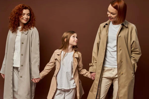 Drei Generationen, fröhliche rothaarige Familie in Mänteln, Händchen haltend vor braunem Hintergrund, Frauen und Mädchen — Stockfoto