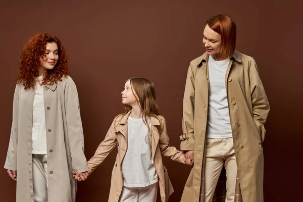 Tres generaciones, alegre familia pelirroja en abrigos tomados de la mano sobre fondo marrón, mujeres y niñas - foto de stock