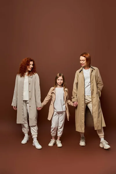 Female generation, joyful redhead family in coats holding hands on brown backdrop, women and girl — Stock Photo